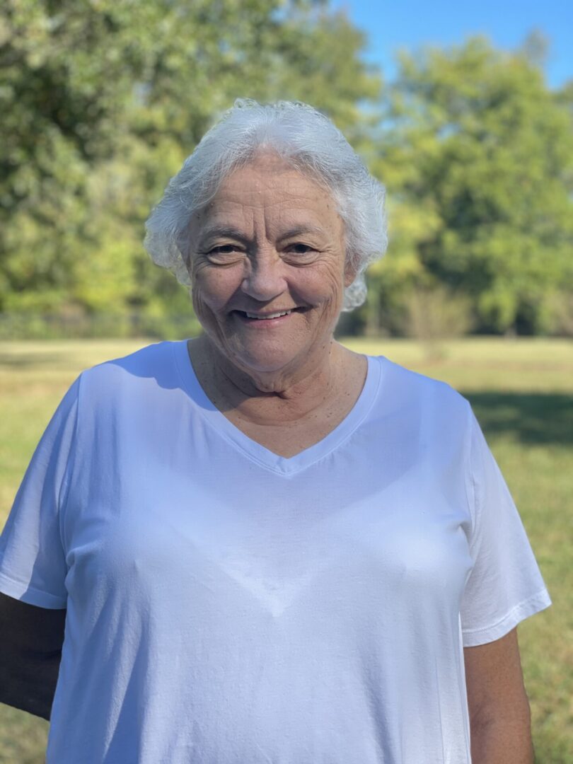 A woman in white shirt standing on grass near trees.