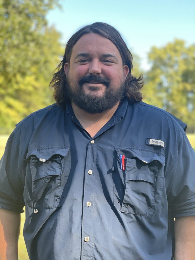 A man with long hair and beard standing in front of trees.